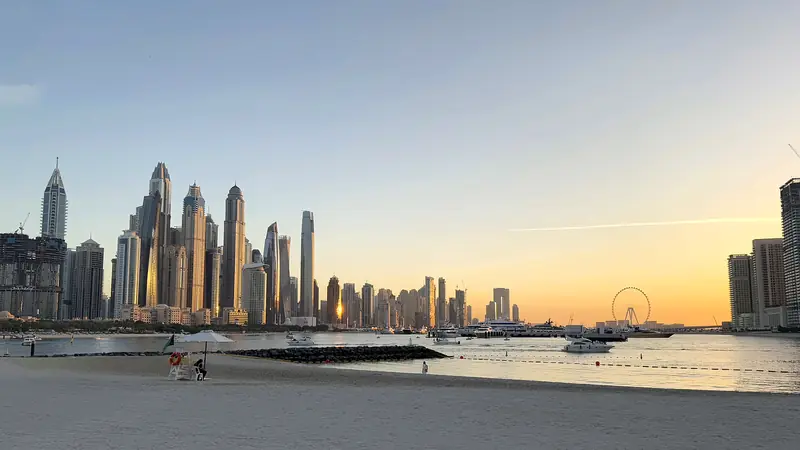 Dubai Marina skyline and the Ain Dubai wheel at sunset, seen across the water from the beach